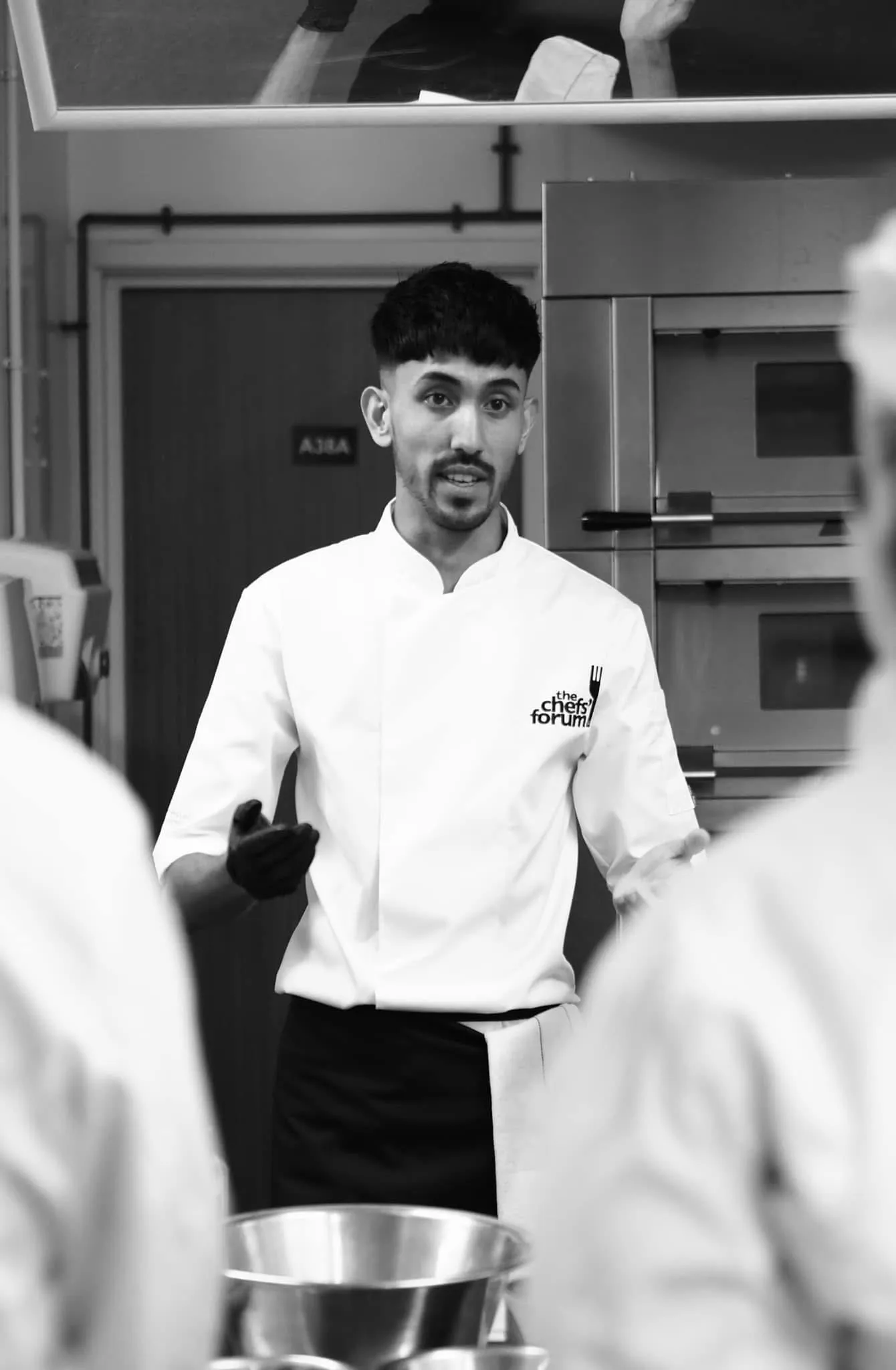 Chef Jubz teaching a class — gloved hand mid-explanation, students' backs visible in the foreground.